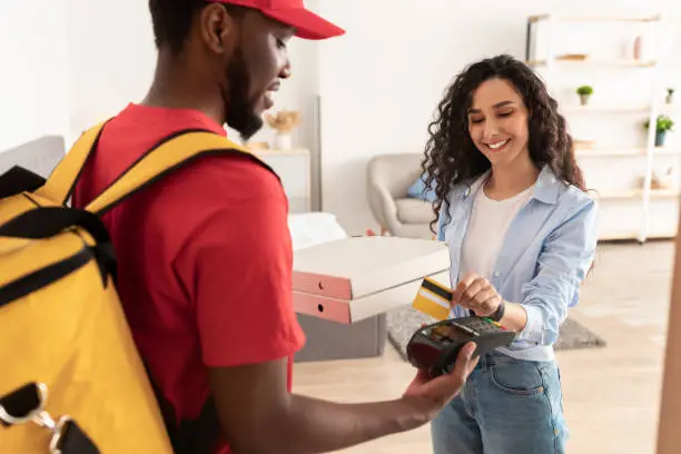 Delivery Concept. Portrait Of Smiling Millennial Female Customer Paying With Credit Card Standing With Courier In Doorway, Receiving Fast Food Pizza Boxes At Home Or Office. Modern Shopping, Shipment
