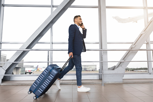 Handsome middle eastern man walking with suitcase and talking on cellphone in airport, young arab businessman carrying luggage and enjoying phone call while going to flight boarding, side view