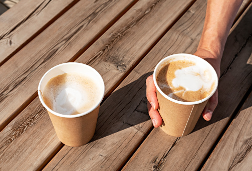 Two cappuccino in beige paper cups on wooden striped table outside and a hand holding one of them, drinking coffee, meeting friends