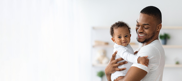 Child Care. Portrait Of Smiling Young Black Father Holding Newborn Baby In Arms While Posing Together Near Window At Home, African Dad Bonding With Cute Infant Kid, Panorama With Copy Space