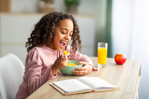 Cute black teen girl having healthy breakfast, eating cereals and drinking orange juice, preparing for lessons and reading book while sitting at desk at home