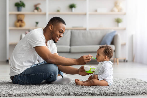 Paternity Leave. Happy Black Dad Feeding Infant Baby From Spoon At Home Paternity Leave. Happy Black Dad Feeding His Infant Baby From Spoon While They Sitting Together On Floor At Home, Young African American Father Taking Care About His Little Newborn Child, Side View paternity stock pictures, royalty-free photos & images