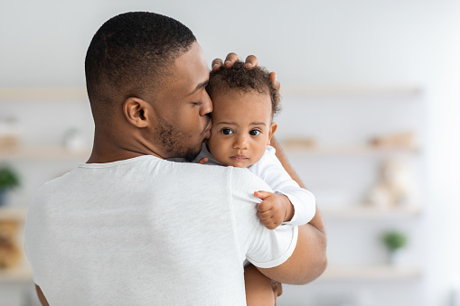 Father's Care. Young Black Dad Holding And Kissing Adorable Newborn Baby At Home, Cute Infant Child Looking At Camera, African American Daddy Bonding With His Son Or Daughter At Home, Closeup Shot