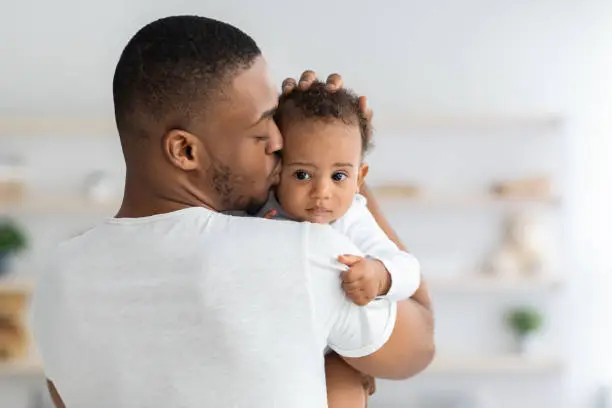 Father's Care. Young Black Dad Holding And Kissing Adorable Newborn Baby At Home, Cute Infant Child Looking At Camera, African American Daddy Bonding With His Son Or Daughter At Home, Closeup Shot