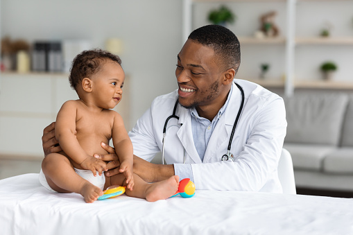 Child's Healthcare Concept. Portrait Of Smiling Black Doctor Making Check Up For Little Infant Baby Boy, Cute Toddler Child In Diaper Having Appointment At Pediatrician's Office, Free Space