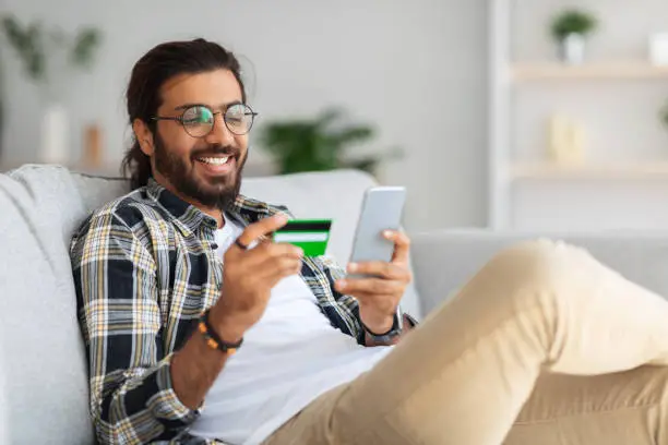 Cheerful young indian man with credit card and mobile phone sitting on couch at home, closeup, copy space. Happy guy freelancer shopping online, using smartphone and holding card, e-commerce