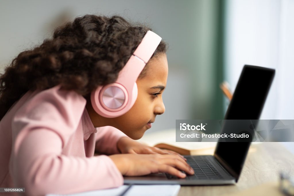 Youth and technology concept. Focused black schoolgirl using laptop computer, sitting at table too close to pc, side view Youth and technology concept. Focused black schoolgirl using laptop computer, sitting at table too close to pc, side view. Little girl with poor eyesight having near vision problem Child Stock Photo