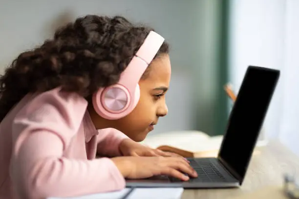 Photo of Youth and technology concept. Focused black schoolgirl using laptop computer, sitting at table too close to pc, side view