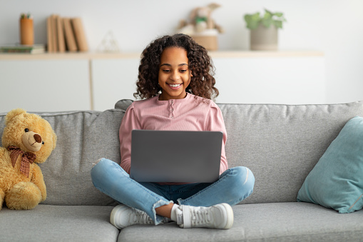 Communication and technology. Smiling black teen girl sitting on sofa and using laptop computer, doing homework, browsing internet, watching video or movie, typing on keyboard and smiling