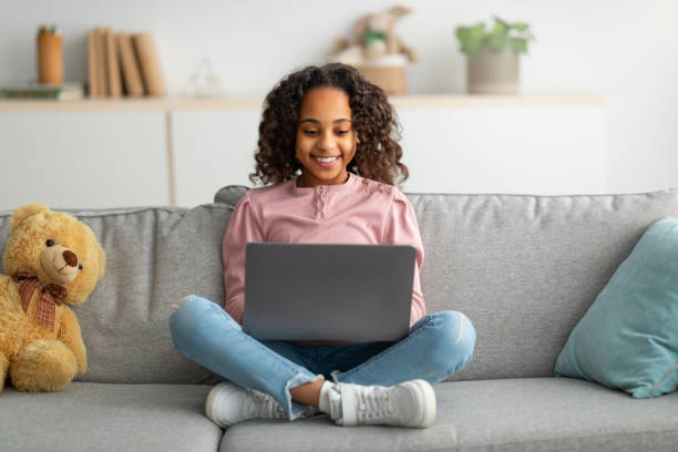 Communication and technology. Smiling black teen girl sitting on sofa and using laptop computer Communication and technology. Smiling black teen girl sitting on sofa and using laptop computer, doing homework, browsing internet, watching video or movie, typing on keyboard and smiling legs crossed at knee stock pictures, royalty-free photos & images