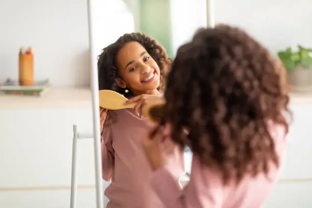 Photo of Morning beauty routine. Happy african american girl combing her curly hair with wooden comb, over shoulder view