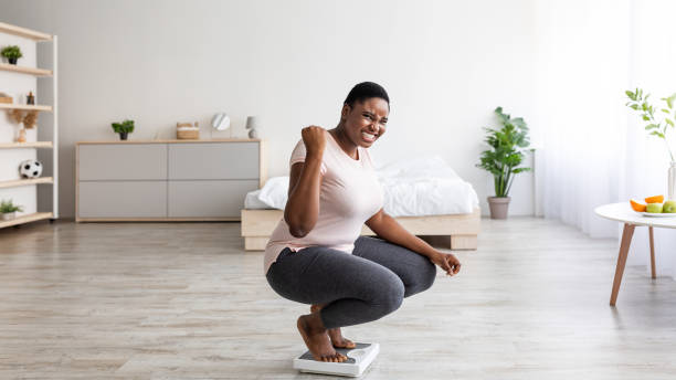 donna nera curvy eccitata seduta su squame a casa, facendo un gesto sì, felice del risultato della sua dieta dimagrante - proporzione foto e immagini stock