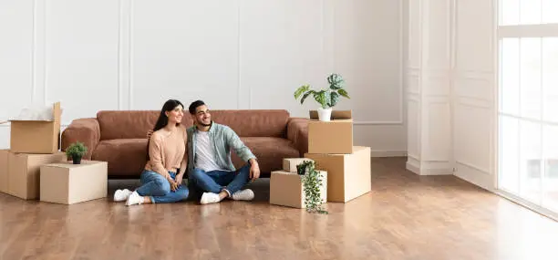 Moving Day. Lovely couple sitting on the floor in new home with cardboard boxes around. Happy man and woman dreaming, looking aside at window, free copy space, panoramic banner for advertisement