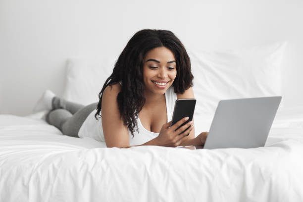 두 단계 인증. 침대에서 스마트 폰과 노트북 컴퓨터를 사용하여 행복한 흑인 여성 - two factor 뉴스 사진 이미지