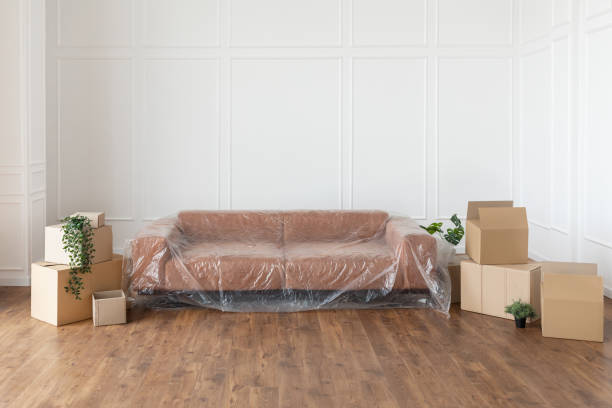 Empty living room with couch, stacks of boxes during relocation Moving Day Concept. Couch under polyethylene cover, cardboard carton boxes stack with household belongings in modern house living room, packed containers on floor in new home. Relocation, renovation belongings stock pictures, royalty-free photos & images