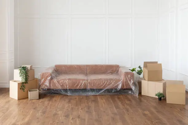 Moving Day Concept. Couch under polyethylene cover, cardboard carton boxes stack with household belongings in modern house living room, packed containers on floor in new home. Relocation, renovation