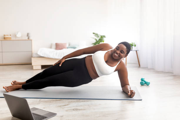 Online sports for weight loss. Overweight black woman doing exercises in front of laptop, standing in side plank at home Online sports for weight loss. Overweight black woman doing exercises in front of laptop, standing in side plank at home. Plus size young lady following video tutorial, training indoors during covid apple core stock pictures, royalty-free photos & images