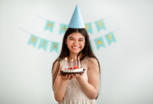 Portrait of Indian teenage girl in festive hat holding birthday cake with candles, smiling at camera indoors. Happy adolescent having party with traditional dessert, celebrating special occasion