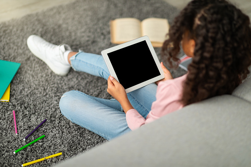 E-learning concept. African american schoolgirl using digital tablet with empty screen, surfing useful website or watching online lesson while sitting on floor with books. Mockup for your design