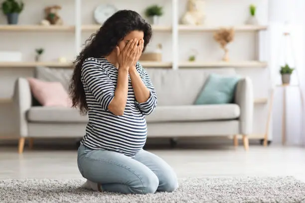 Pregnancy hormonal changes concept. Young curly pregnant woman feeling down, sitting on floor in living room and crying, covering her face with both palms, staying alone at home, free space