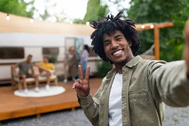 Photo of Black guy taking selfie near motorhome, gesturing peace, resting with his multiracial friends outdoors, enjoying summer