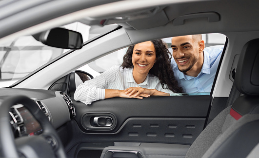 Cheerful young arab man and woman buying new car, checking automobile salon through the window. Smiling middle-eastern couple looking inside beautiful black auto in showroom, purchasing auto