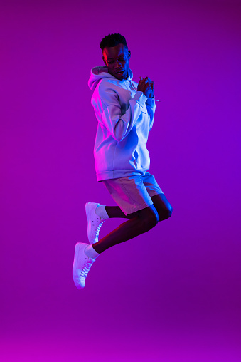 Full lenght portrait of young energetic African American man jumping in dark purple futuristic cyberpunk neon light background