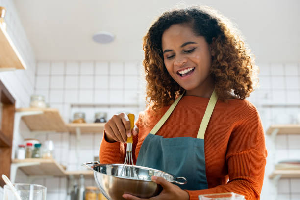 junge hübsche afroamerikanerin, die sich beim kochen in der küche amüsiert, während sie zu hause bleibt - rühren stock-fotos und bilder