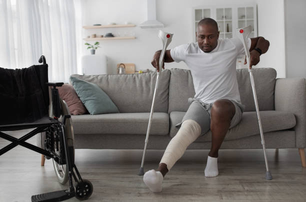Bone fracture after incident, treatment at home, recovery Bone fracture after incident, treatment at home, recovery. Black middle aged male with broken leg with plaster cast gets up from sofa with crutches in living room interior with wheelchair, empty space spinal surgery recovery stock pictures, royalty-free photos & images