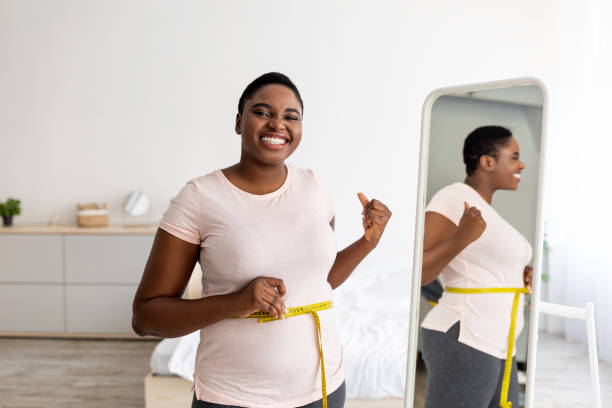 mujer negra de talla grande que mide la cintura con cinta adhesiva frente al espejo, mostrando los resultados de la dieta adelgazante, gesticulando el pulgar hacia arriba - régimen alimenticio fotografías e imágenes de stock