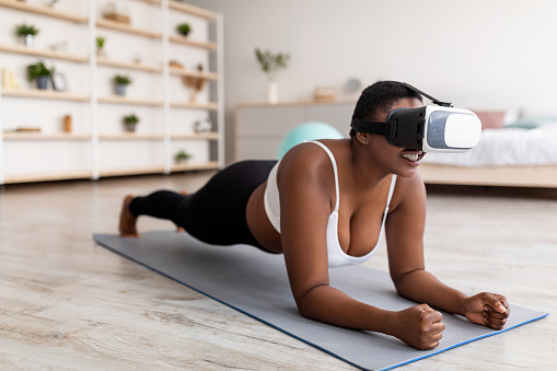 Domestic virtual training. Curvy young black lady in VR glasses standing elbow plank at home. Overweight millennial woman training via augmented reality app. Sports and technologies concept