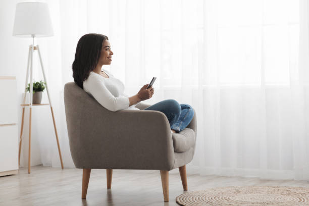 istock Relaxed black lady sitting in armchair and using smartphone at home, checking email, side view, copy space 1331265080