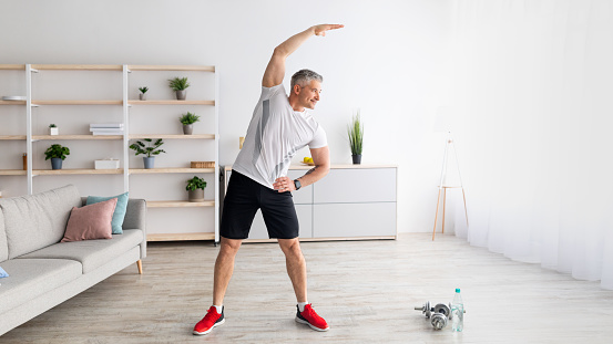Sport on retirement. Active mature man doing flexibility exercises for healthier living during home workout. Man leading sporty lifestyle, training in living room interior, copy space, panorama