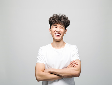 Portrait of happy young man wearing white t-shirt. Male student standing with arms crossed and laughing with eyes closed. Studio shot, grey background.