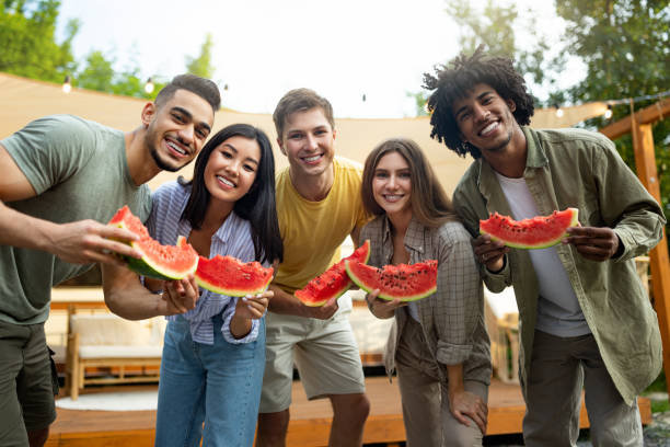 Portrait of cheerful multiracial friends holding pieces of watermelon, smiling at camera near camper van outdoors Portrait of cheerful multiracial friends holding pieces of watermelon, smiling at camera near camper van outdoors. Millennial diverse youth spending summer holidays on camping trip black man eating watermelon stock pictures, royalty-free photos & images