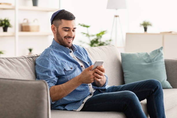istock Smiling jew man using mobile phone sitting on comfy couch 1331267896