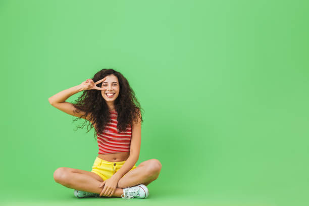 Image of young woman 20s rejoicing and smiling while sitting on floor with legs crossed Image of young woman 20s rejoicing and smiling while sitting on floor with legs crossed against green wall legs crossed at knee stock pictures, royalty-free photos & images