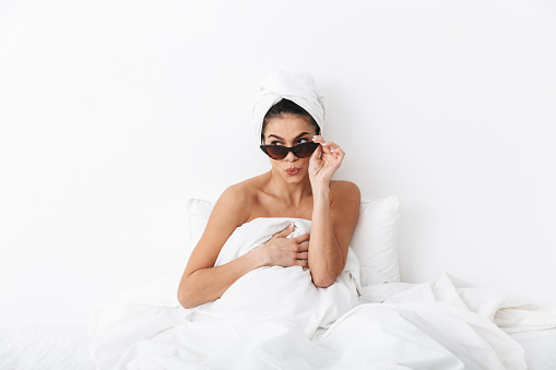 Cheerful young woman sitting in bed after shower wrapped in blanket, wearing sunglasses