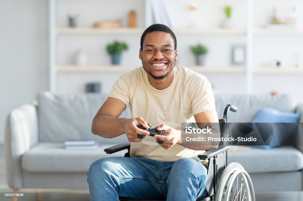 Cheerful disabled man in wheelchair enjoying videogame on playstation, having fun at home Cheerful disabled man in wheelchair enjoying videogame on playstation, having fun at home. Handicapped male gamer with joystick playing computer arcade. Quarantine hobbies concept Video Game Stock Photo