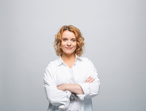 Portrait of young woman with red curly hair wearing white shirt. Female student standing with arms crossed and smiling at camera. Studio shot, grey background.