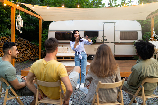 Fun outdoor entertainments. Happy Asian woman playing word guessing game with multiracial friends near RV at campsite. Group of cheerful young people enjoying charades or pantomime riddles, outdoors