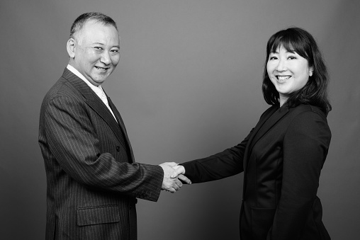 Studio shot of mature Asian businessman and mature Asian businesswoman together against gray background in black and white
