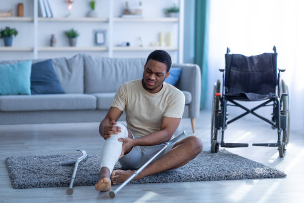 Black man with injured leg sitting on floor after falling down, having trouble walking with crutches next to wheelchair Young black man with injured leg sitting on floor after falling down, having trouble walking with crutches, wheelchair standing nearby. African American guy using mobility aid with no success crutch stock pictures, royalty-free photos & images