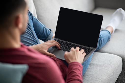 Arab guy using modern laptop with empty screen at home, mockup. Unrecognizable middle-eastern millennial man reclining on couch and using notebook with blank display, over shoulder shot