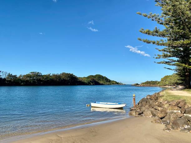 River Coastline with Row Boat Moored Near Sand Horizontal seascape of still calm river waters with small row boat dinghy near sand with pine tree lined coastline horizon going out to sea at Brunswick Heads River near Byron Bay NSW Australia sailing dinghy stock pictures, royalty-free photos & images