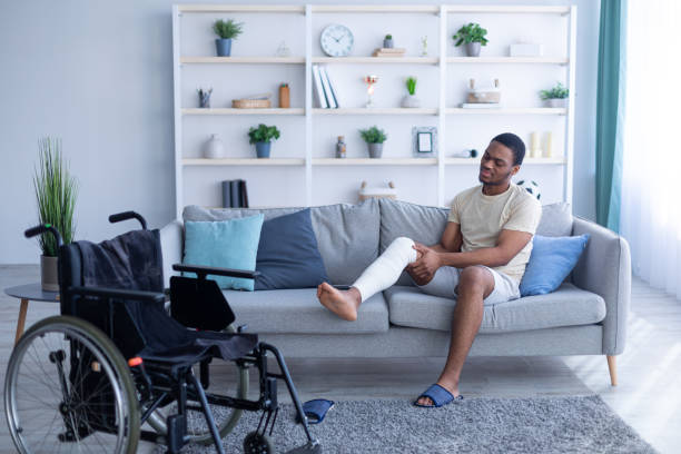 Young black guy with injured leg sitting on sofa, trying to get to wheelchair at home, full length Young black guy with injured leg sitting on sofa, trying to get to wheelchair at home, full length. Millennial man suffering from pain in plastered limb, having trouble moving around house, copy space orthopedic cast stock pictures, royalty-free photos & images