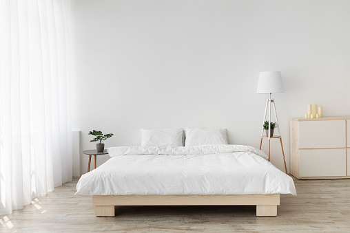 Ideas for scandinavian minimalist bedroom. Double bed with pillows, soft white blanket, lamp and furniture on wooden floor. White wall, big window with curtains in bedroom interior