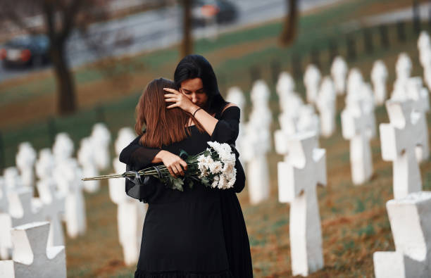서로를 껴안고 울고 있습니다. 검은 옷을 입은 두 명의 젊은 여성이 많은 흰색 십자가를 가진 묘지를 방문합니다. 장례식과 죽음의 개념 - mourner 뉴스 사진 이미지