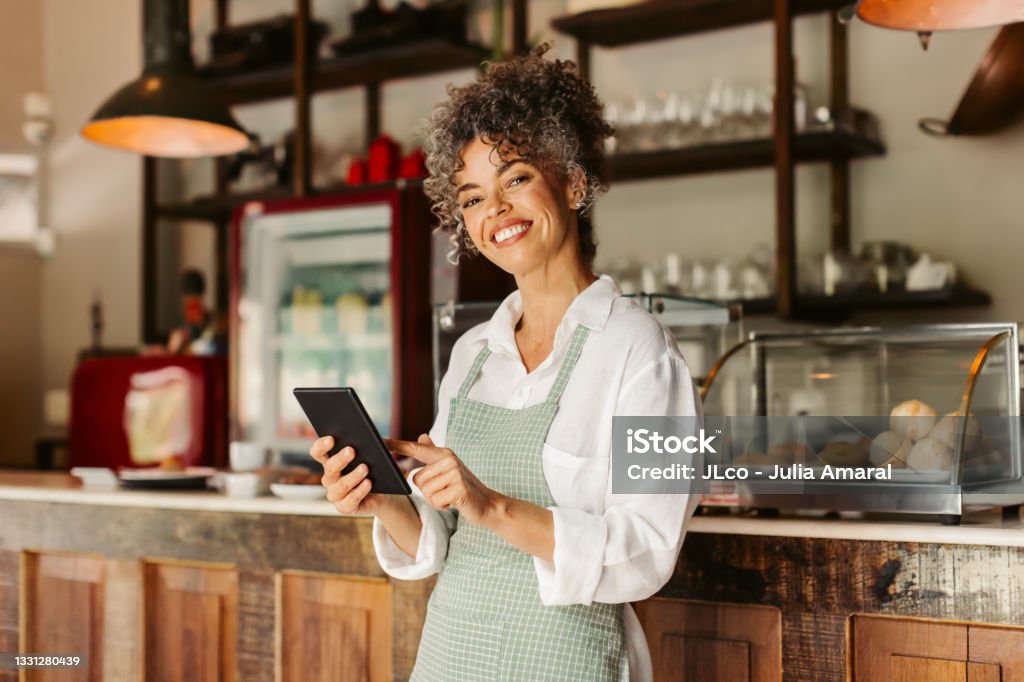 Smiling entrepreneur holding a digital tablet in her cafe Entrepreneur holding a digital tablet in her cafe. Mature female cafe owner smiling at the camera while standing in front of the counter. Successful businesswoman running her business online. Occupation Stock Photo