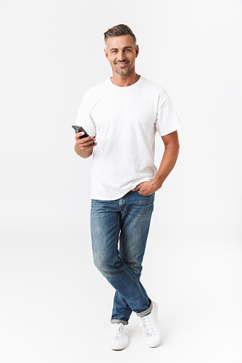 Full length image of muscular man 30s wearing casual t-shirt and jeans using mobile phone while holding in hand isolated over white background